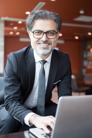 Closeup portrait of smiling middle-aged handsome business man looking at camera and working on laptop computer in lobby